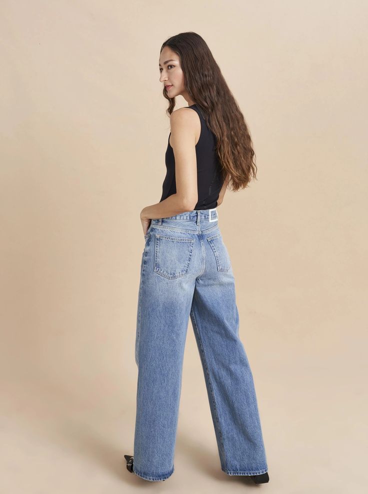 a woman wearing high rise jeans and black tank top standing in front of a beige background