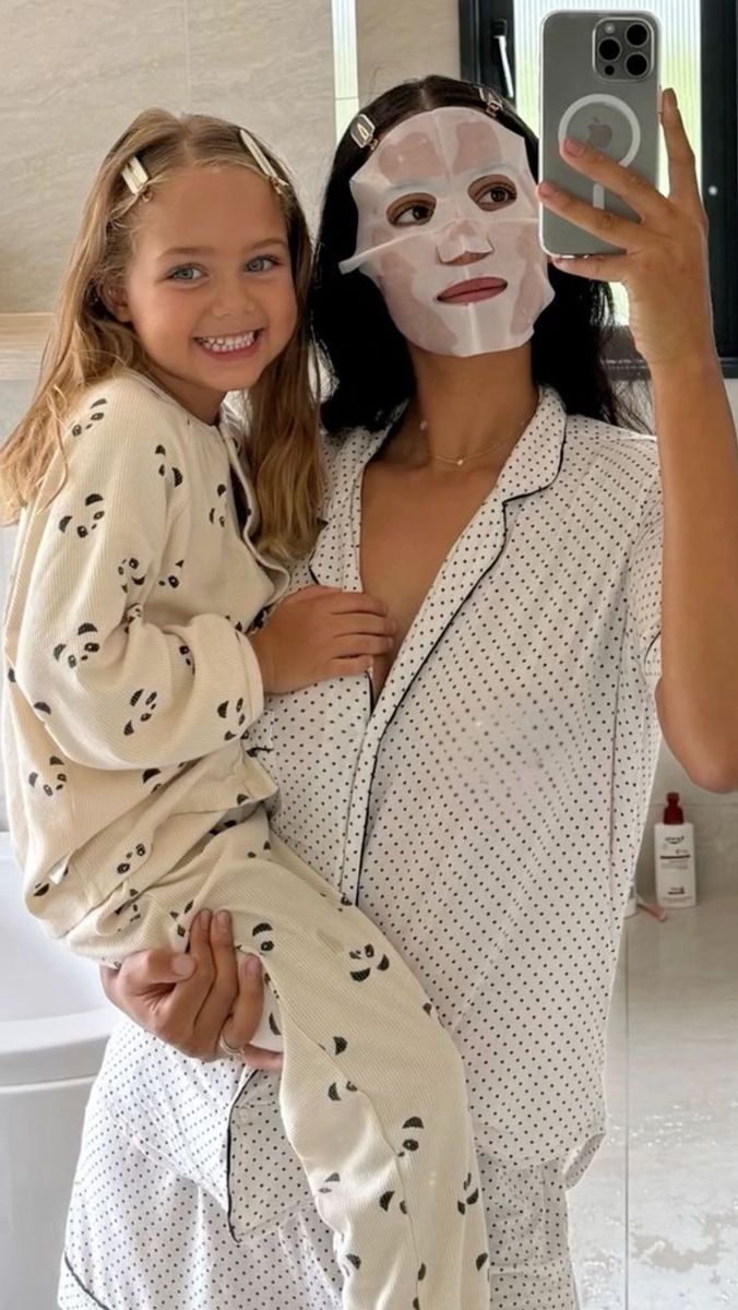 a woman in pajamas holding a child wearing a face mask and taking a selfie