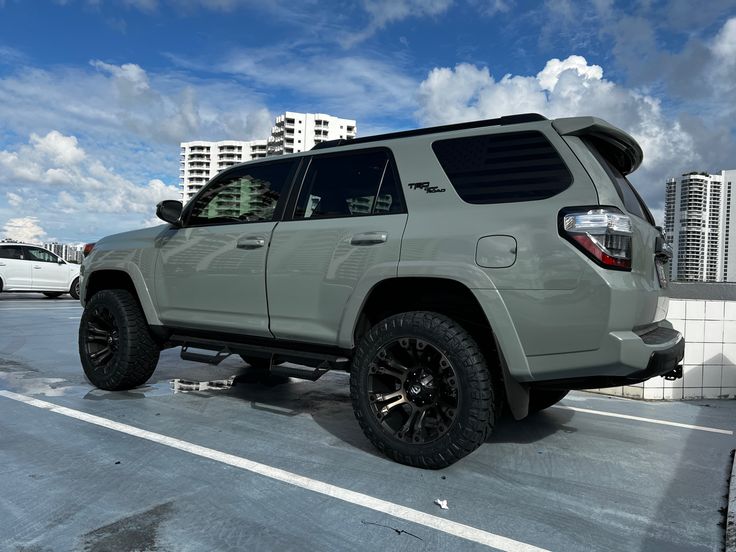 a gray four door suv parked in a parking lot next to tall buildings and clouds