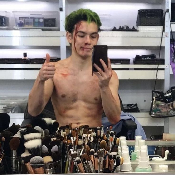 a man with green hair is holding up his cell phone and surrounded by makeup brushes