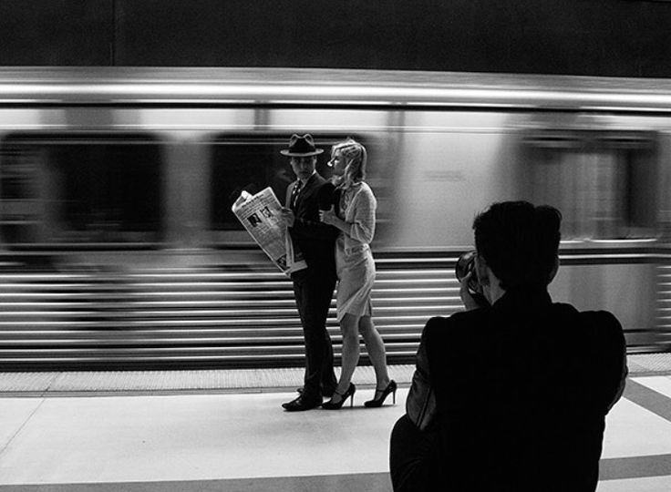 two people are standing on the platform as a train passes by in black and white