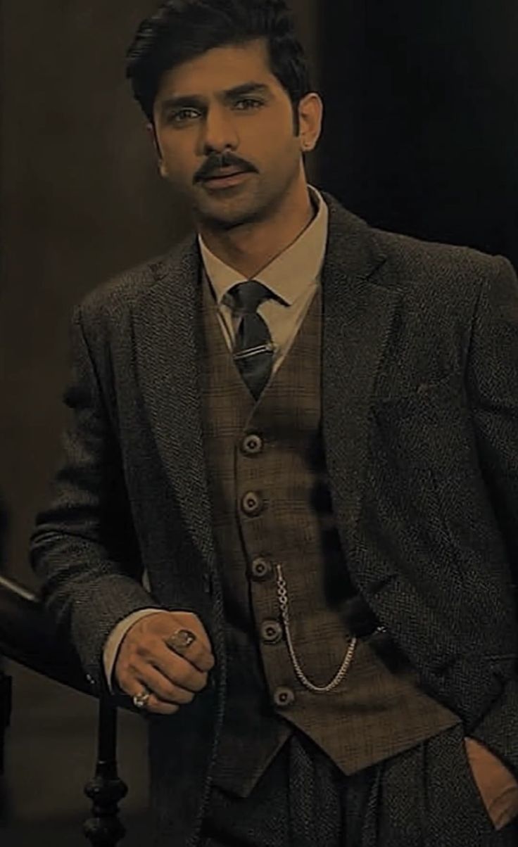 a man wearing a suit and tie standing in front of a staircase with his hands on his hips