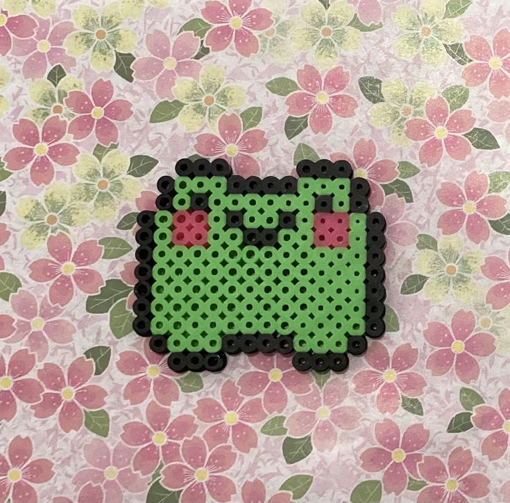 a green piece of art made out of perler beads on a pink flowered background