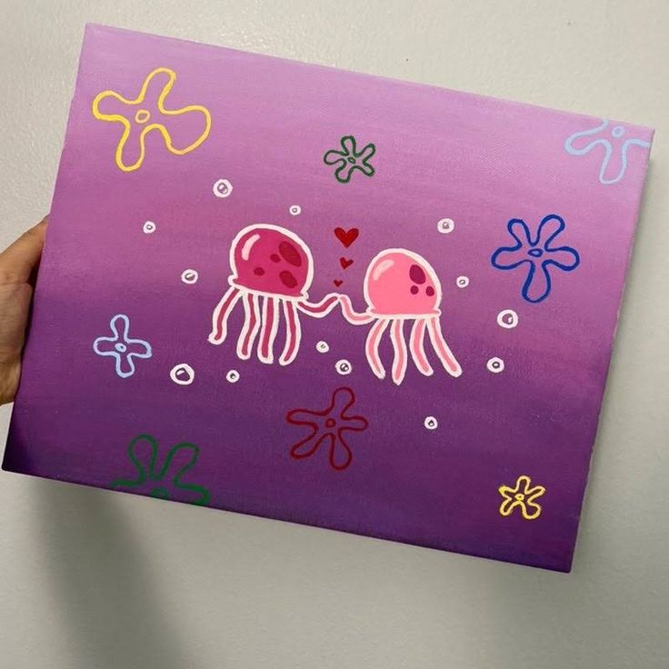 a hand holding up a purple painting with two jellyfishs on it's sides