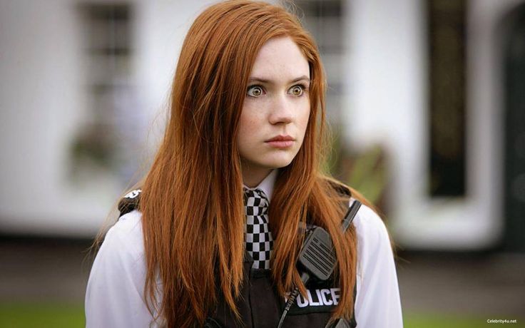 a woman with long red hair wearing a police vest and checkered tie standing in front of a house