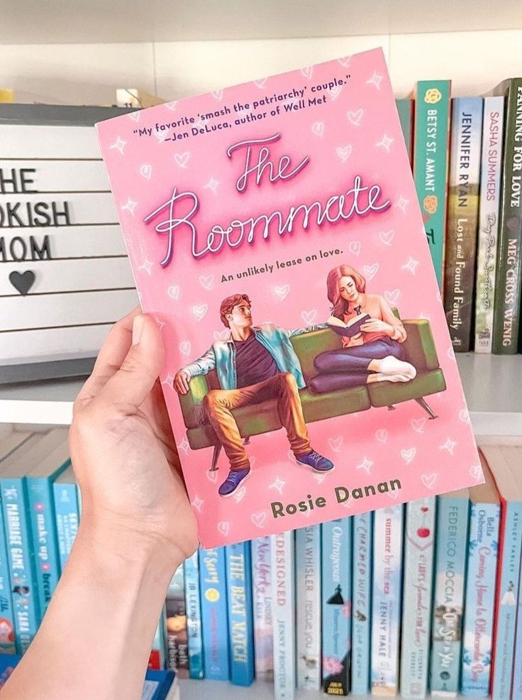 a person holding up a pink book in front of a shelf with books on it