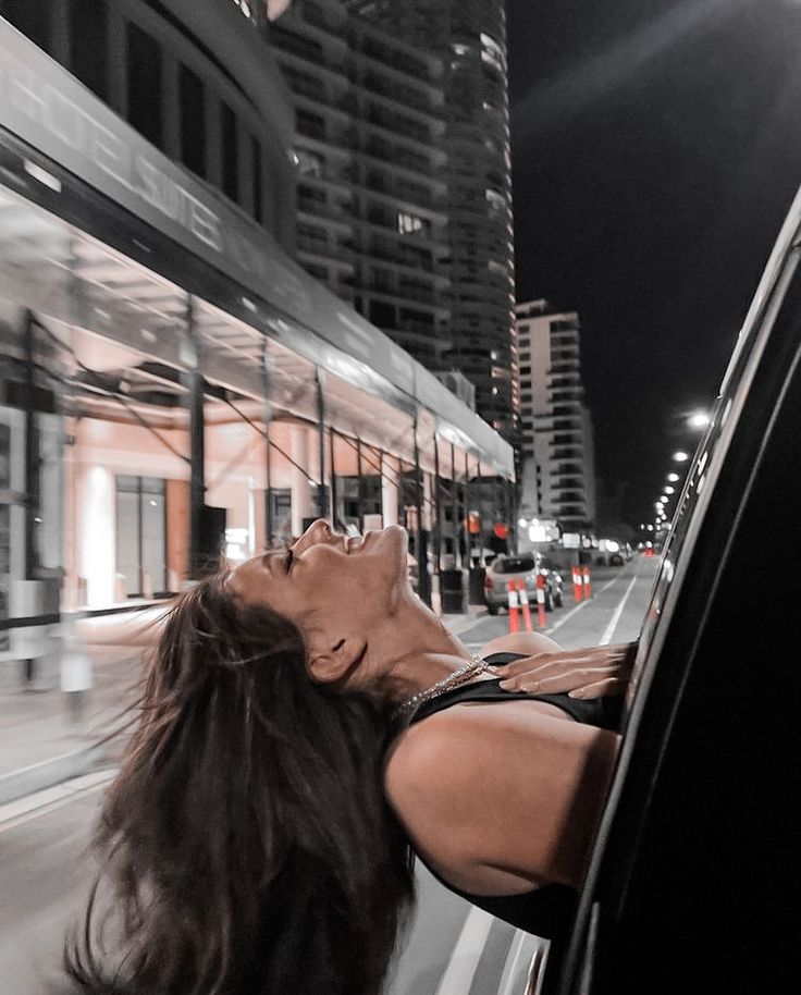 a woman leaning out the window of a car on a city street at night with buildings in the background