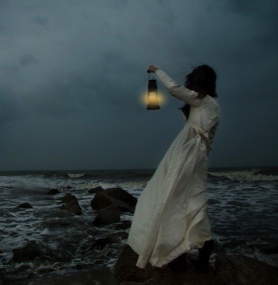 a woman in white dress standing on rocks near the ocean holding a light up lantern