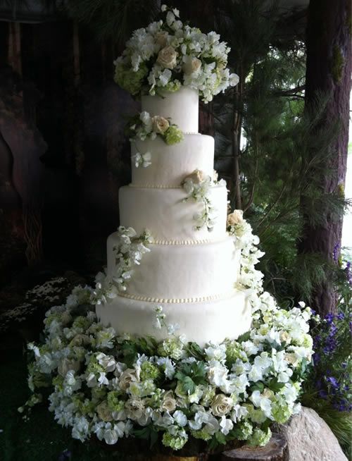 a wedding cake with white flowers on top and greenery around the bottom, in front of a window