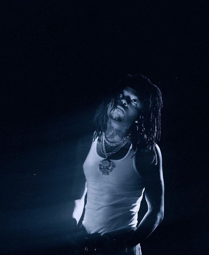 a man with dreadlocks standing in the dark wearing a white tank top and black pants