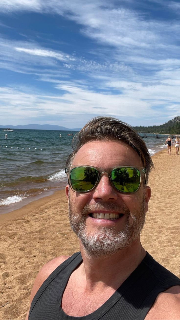 a man wearing sunglasses standing on top of a sandy beach next to the ocean with people in the background