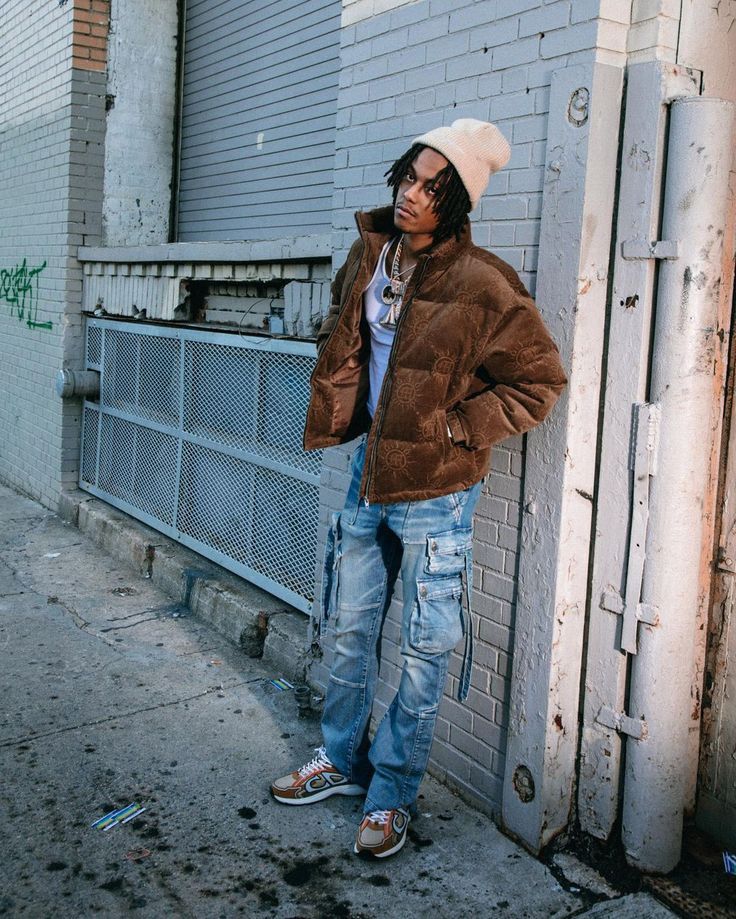 a man leaning against a wall wearing a jacket and hat with his hands in his pockets