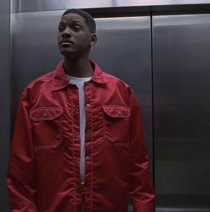 a man wearing a red jacket standing in front of a metal wall with his hands in his pockets