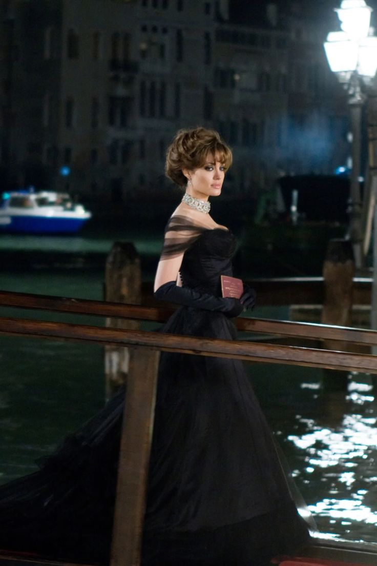 a woman in a black dress is standing on a pier at night with her hands behind her back