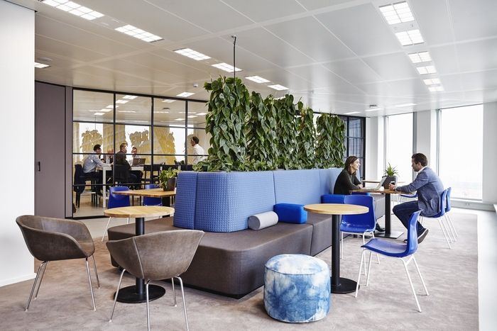 people are sitting at tables in an office with plants on the wall behind them and blue couches