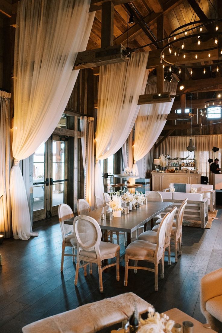 A spacious rustic-chic wedding reception space with a draped ceiling, exposed wooden beams, and warm lighting. A long dining table with neutral tones and natural elements creates a cozy and elegant atmosphere. Bridal Suite Design, Wedding Reception Draping, Barn Reception Decorations, Draping Wedding Reception, Classy Barn Wedding, Glam Wedding Reception, Draped Ceiling, Neutral Wedding Palette, Rustic Barn Wedding Reception