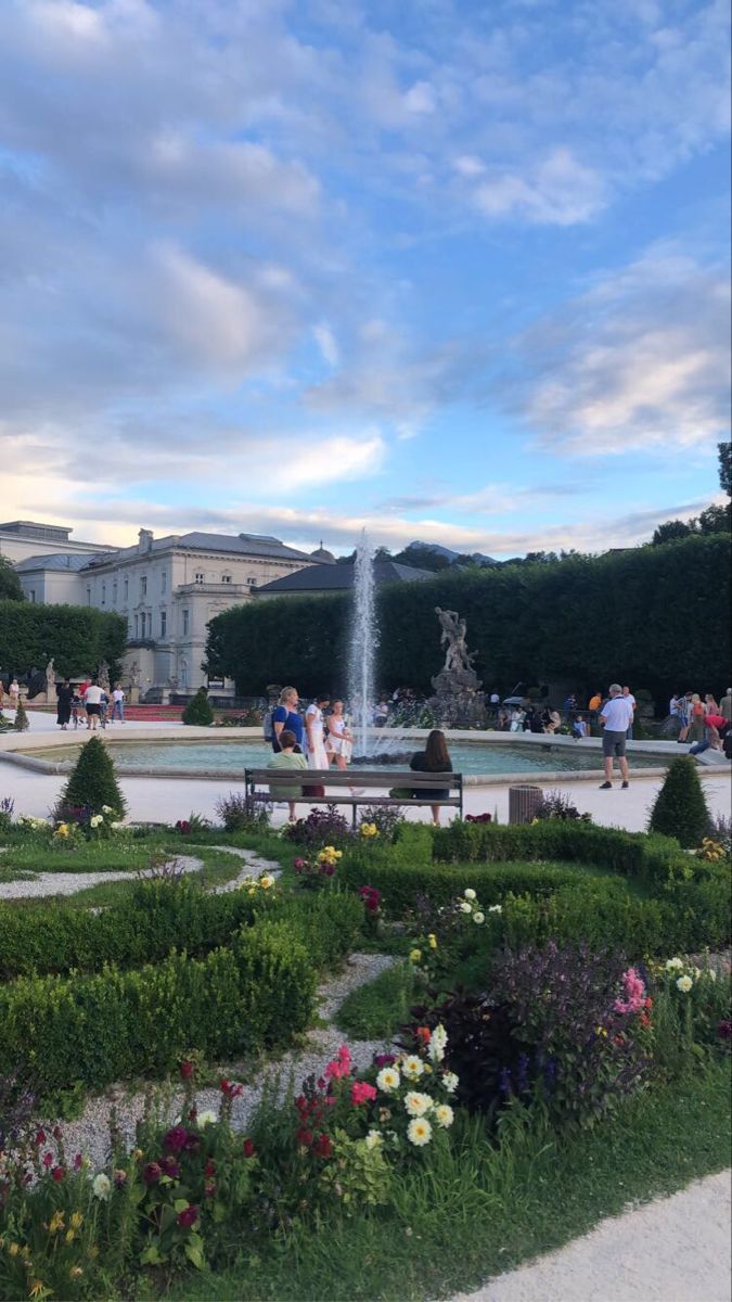 a fountain surrounded by flowers and greenery in front of a building with people walking around it