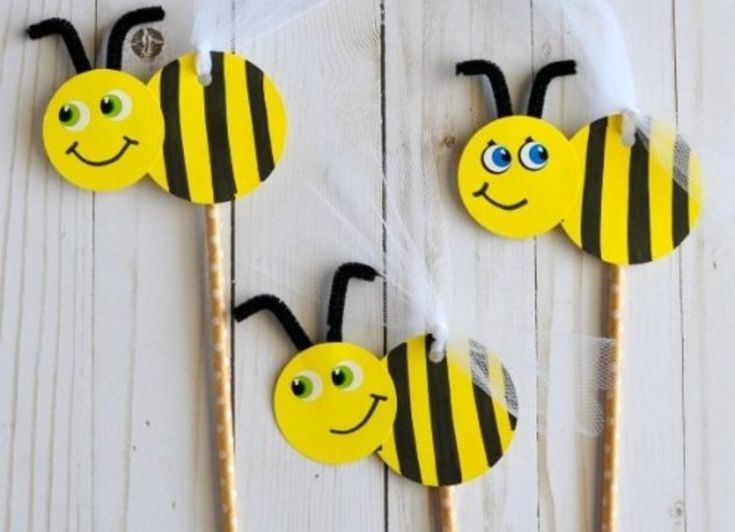 three bees on sticks with faces drawn on them, sitting on a white wooden surface