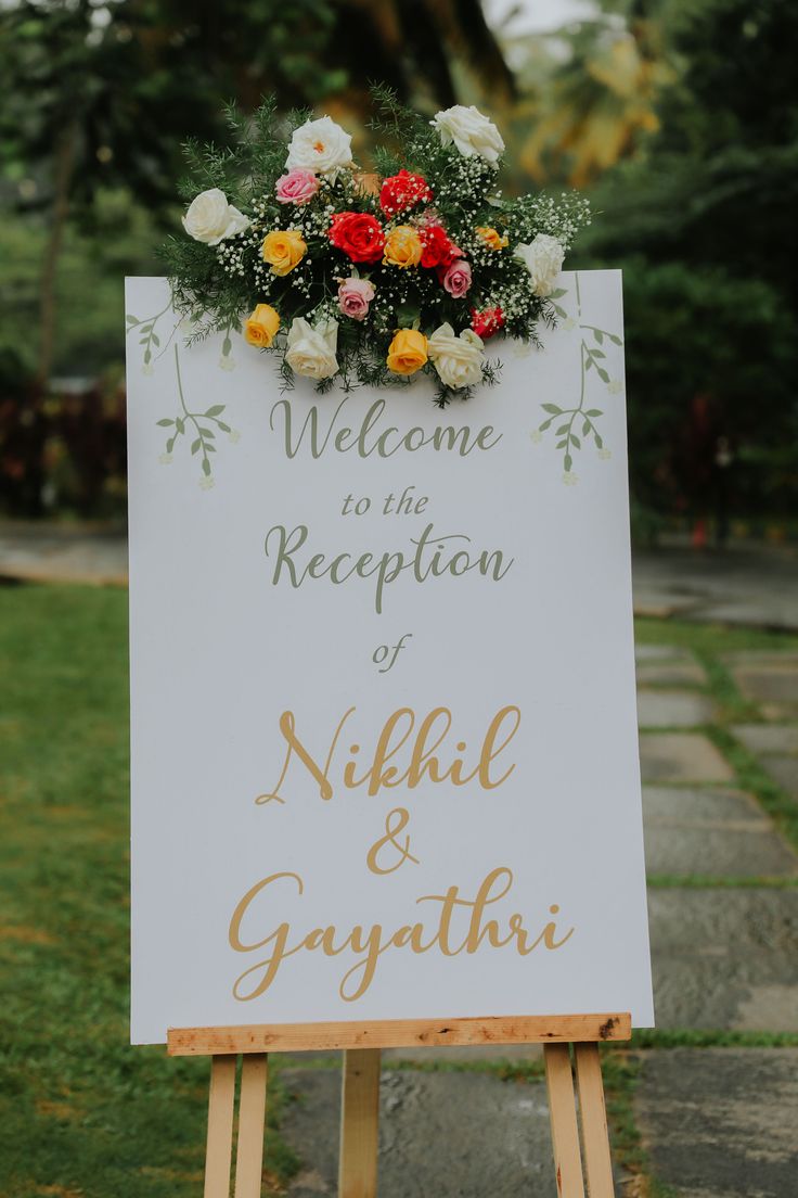 a welcome sign with flowers on it in front of some grass and trees at an outdoor venue