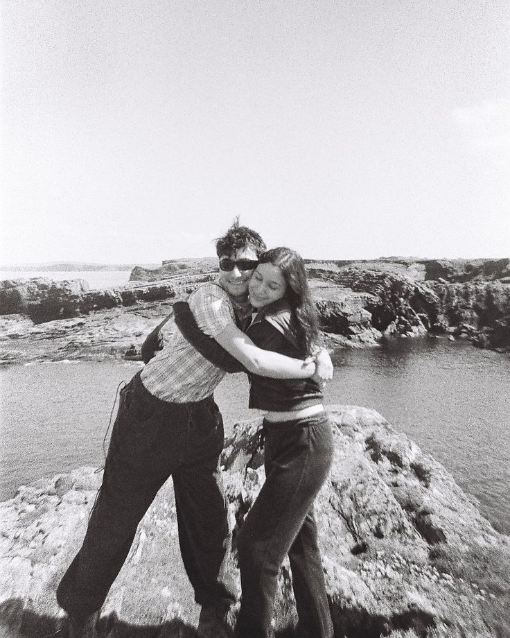 two people standing on top of a rock near the ocean and one person is hugging