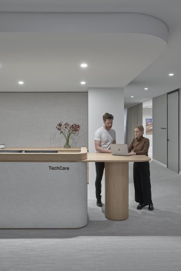 two people standing in an office with a laptop on the desk and another person looking at it