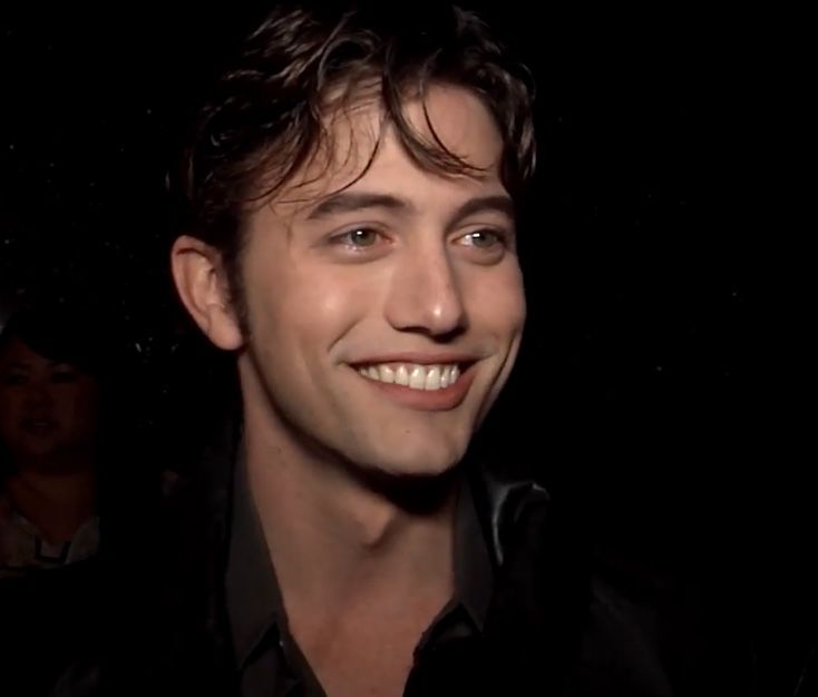 a man smiling and wearing a black shirt in front of a dark room with other people