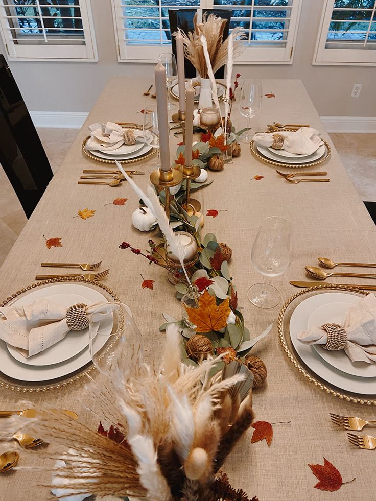 the table is set for thanksgiving dinner with white and gold plates, silverware, and fall leaves