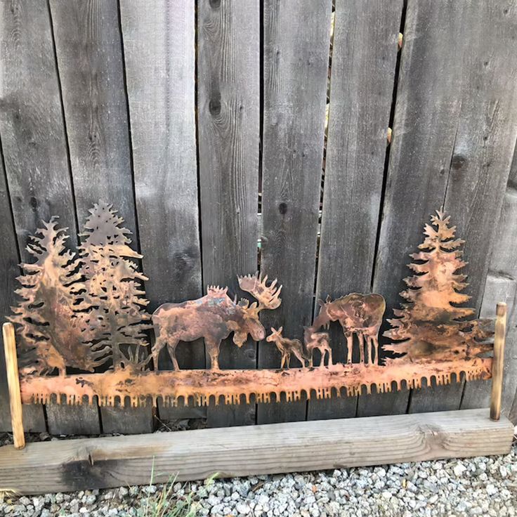 three metal moose sculptures sitting on top of a wooden bench next to a fence with pine trees