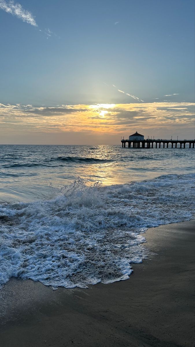 the sun is setting over the ocean with waves crashing on the shore and a pier in the distance