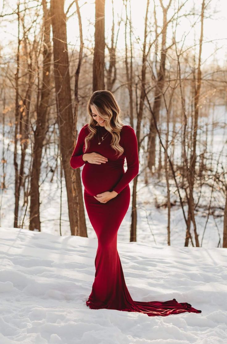 a pregnant woman standing in the snow wearing a long red dress and holding her belly