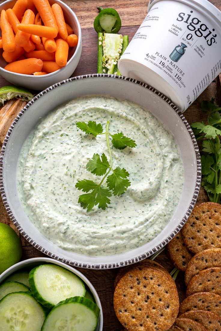 a white bowl filled with dip surrounded by crackers, cucumbers and carrots