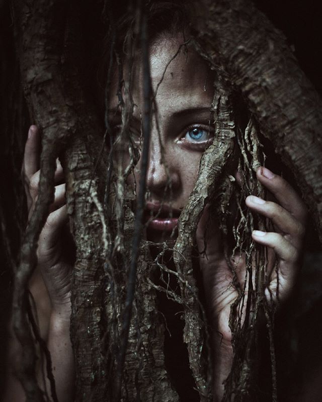 a woman with blue eyes is hiding behind a tree in the dark, holding her hands up to her face