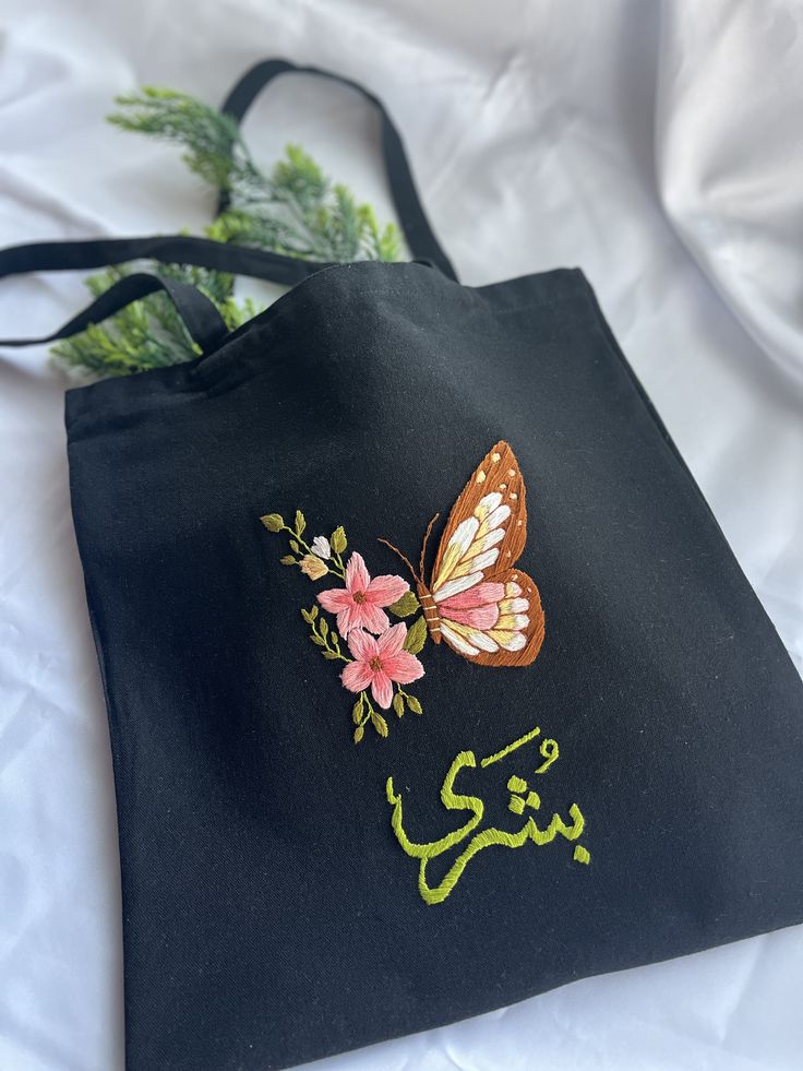 a black tote bag with an embroidered butterfly and flowers on the side, sitting on a white sheet