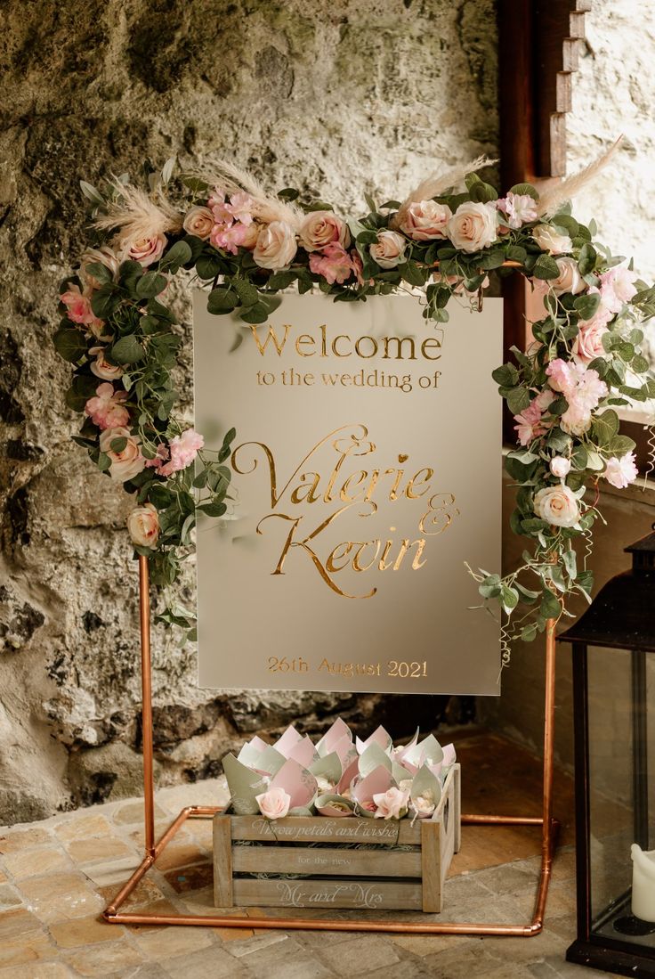 a welcome sign with pink flowers and greenery is displayed in front of a stone wall