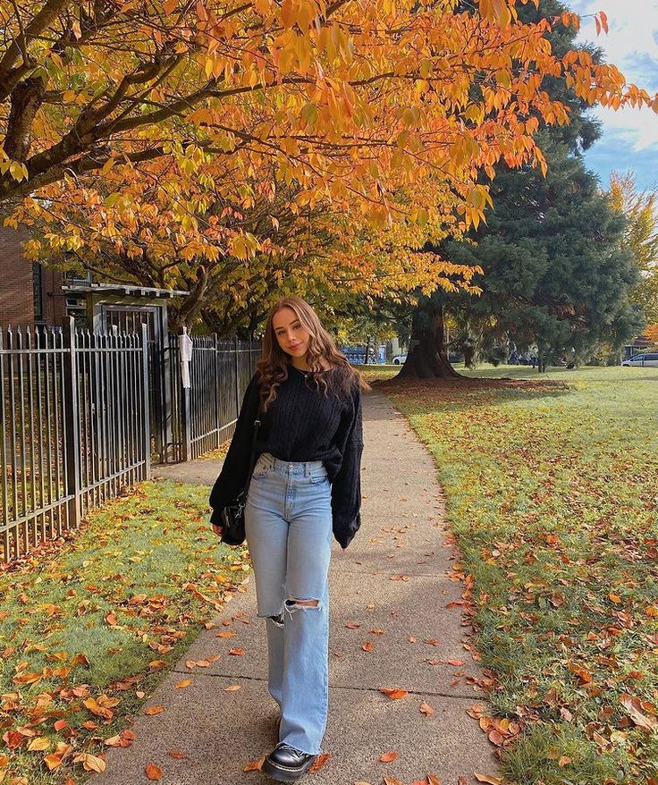 a woman walking down a sidewalk in front of trees with leaves on the ground and grass