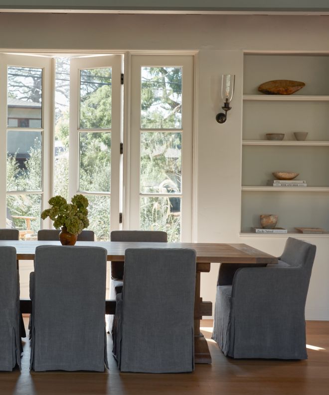 a dining room table with chairs and a vase filled with flowers in front of two large windows