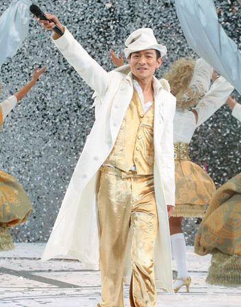 a man in gold pants and white jacket holding his arms up while dancing with other people behind him