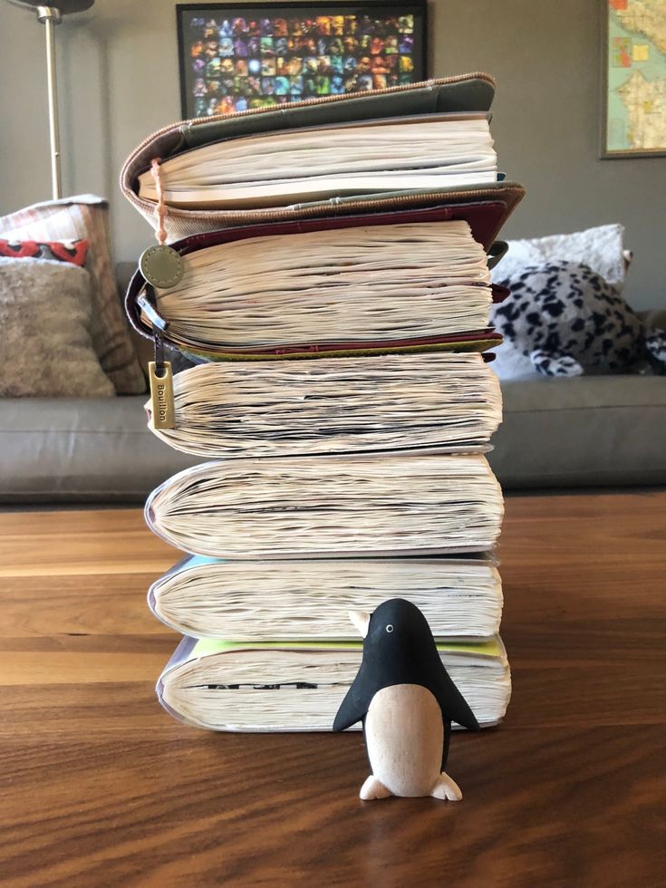 a stack of books sitting on top of a wooden table next to a toy penguin