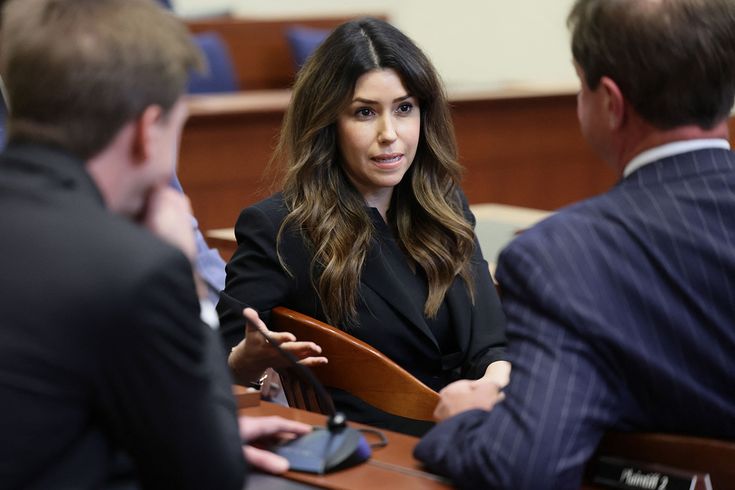 a woman sitting at a table talking to two other people in a room with wooden chairs