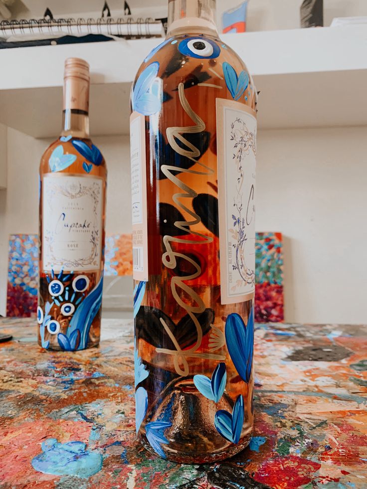 two bottles of alcohol sitting on top of a table covered in colorful paint and paper