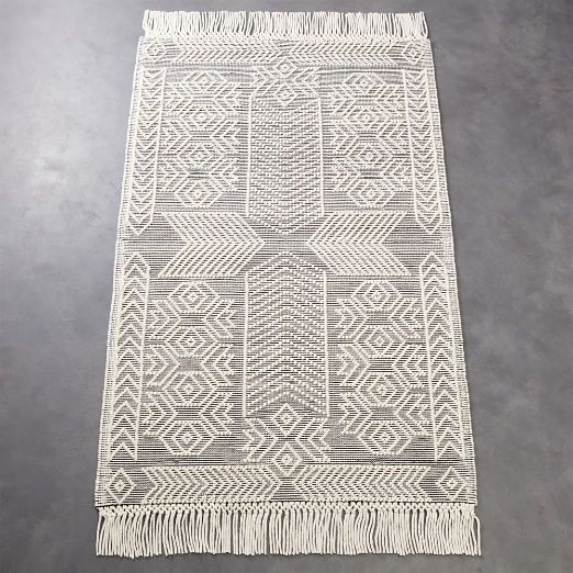 a white and grey rug with fringes on top of the floor in front of a gray wall