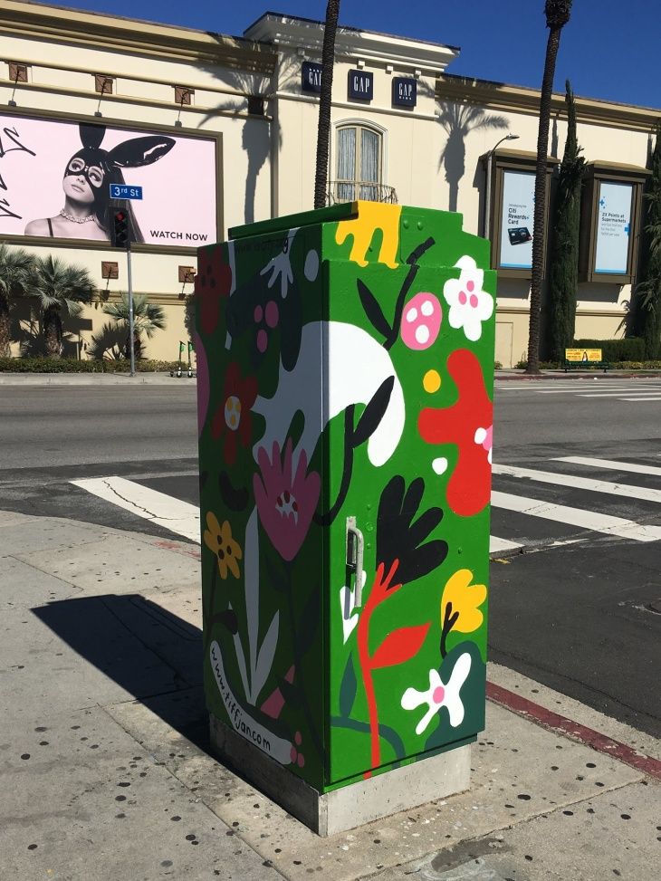 a green box with flowers painted on it sitting on the side of a road next to a building