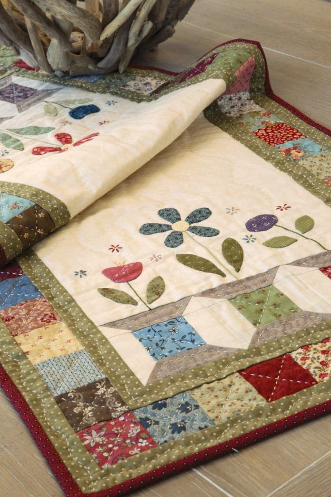 a quilted table runner on the floor next to a vase with flowers in it