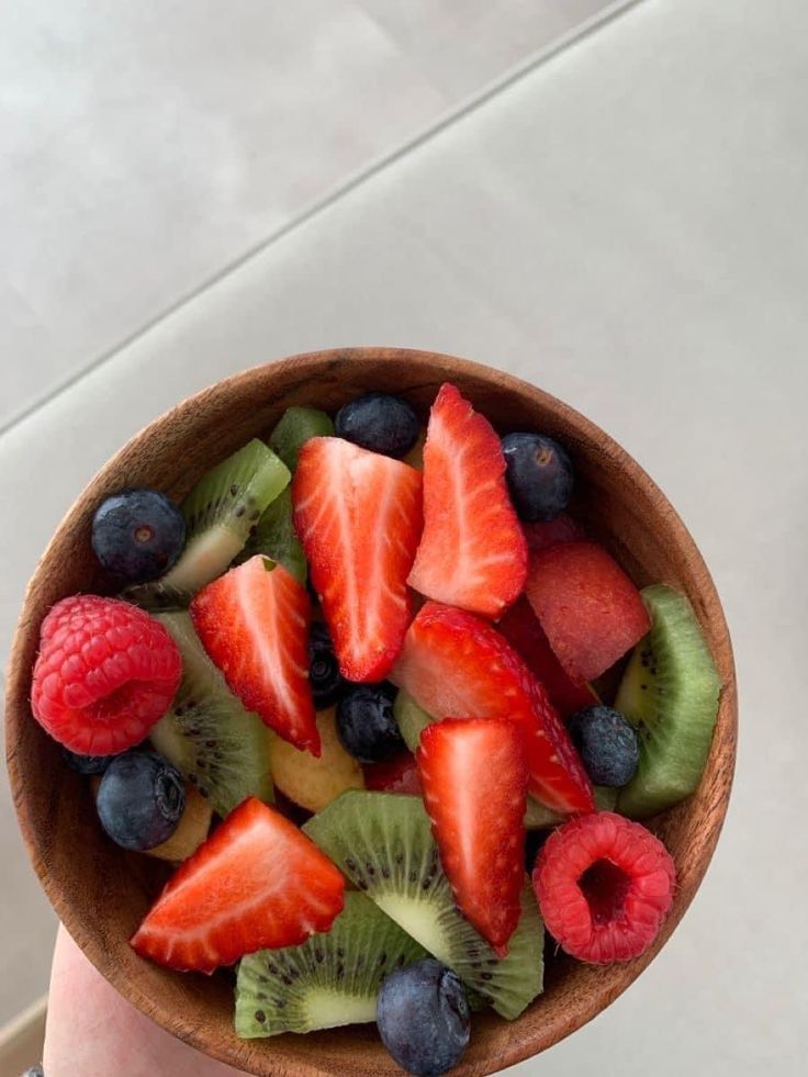 a person holding a wooden bowl filled with sliced up kiwis and strawberries