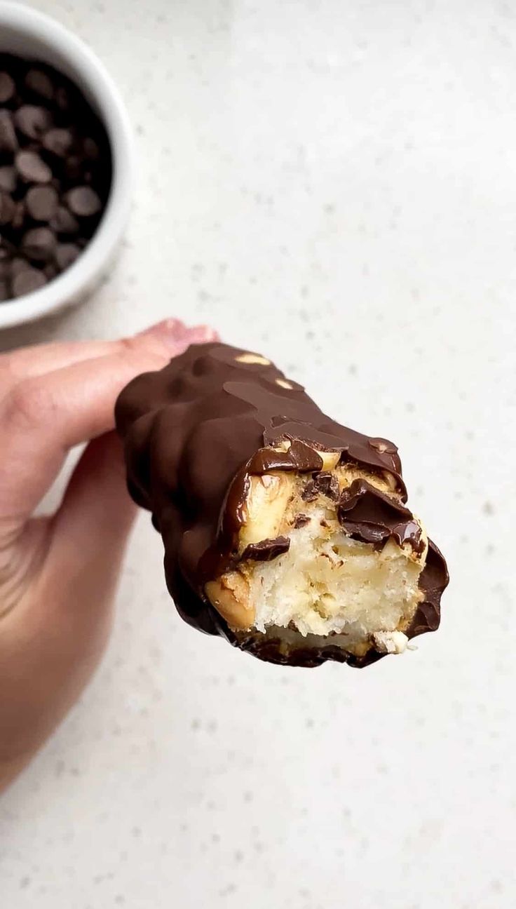 a hand holding a half eaten chocolate covered doughnut next to a bowl of chocolate chips