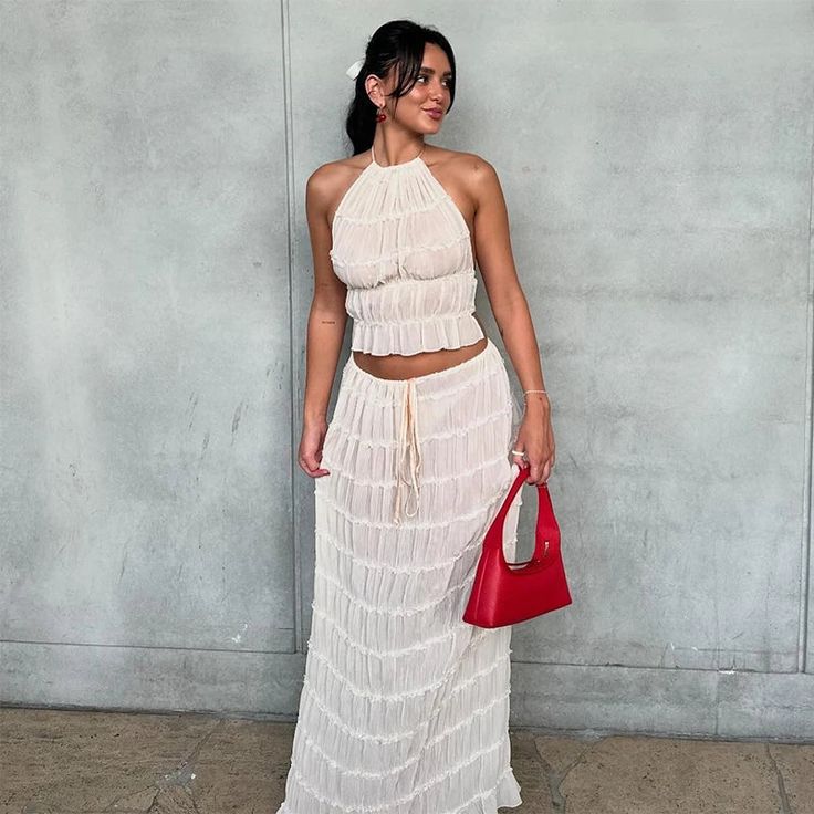 a woman standing in front of a wall holding a red purse and wearing a white dress