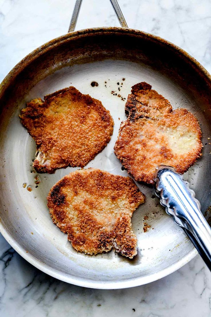 three fried meat patties in a frying pan with tongs on the side