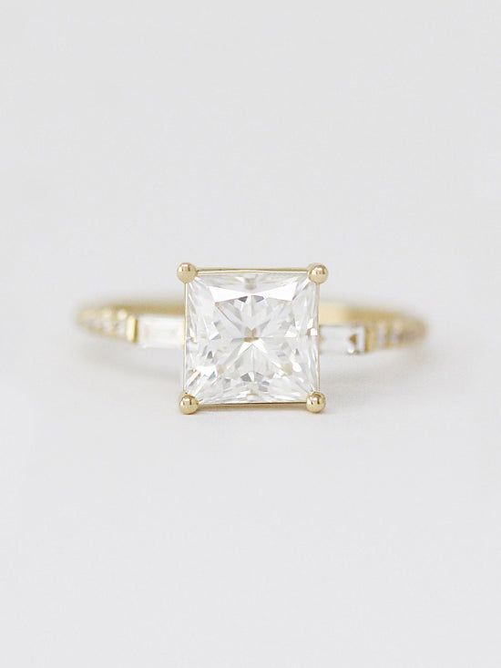 a gold ring with a princess cut diamond in the center, on a white background