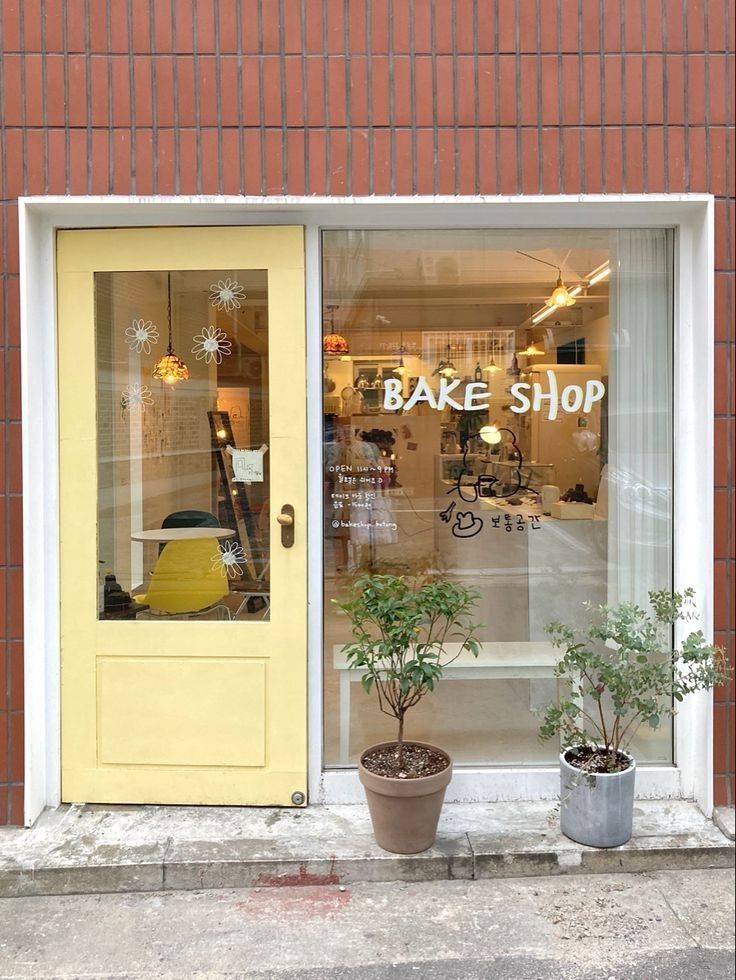 two potted plants sit outside the window of a bake shop with yellow doors