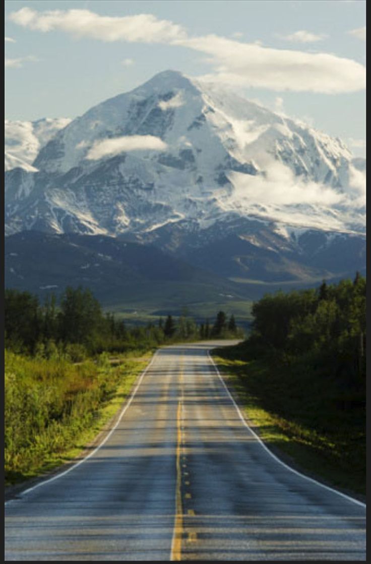 an empty road in front of a snow covered mountain with trees and grass on both sides